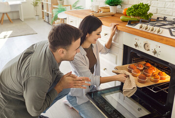 Happy smiling couple pulling baked muffins from oven in kitchen at home together. Young man and woman cooking tasty dessert preparing bakery indoors. Home leisure, food and relationships concept.