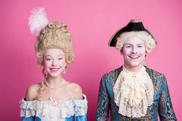 Young couple in baroque costumes posing against a vibrant pink background, exuding charm and...