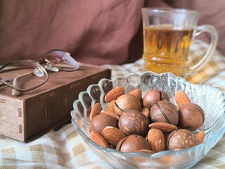 macadamia and almond in glass transparent bowl, glasses and cup of tea on brown background