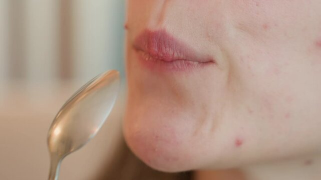 Elegant young lady dips tablespoon of hot chocolate drink into mouth, slowly bringing it out with satisfied expression, focus on lips and spoon with soft indoor background in warm lighting ambiance