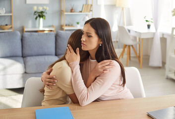 Loving mother hugging and comforting upset teenage daughter at table in living room. Worried parent...