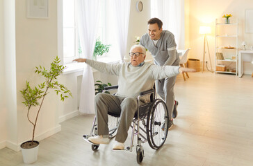 Fototapeta premium Male caregiver having fun together with his senior man patient, riding him in wheelchair, taking care in nursery room, giving help and entertaining. Old people care and assistance concept