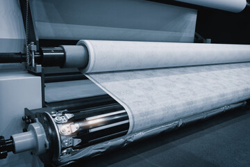 A machine is rolling out a white fabric. The machine is in a factory. The fabric is being made in a...