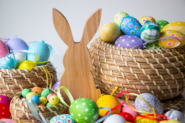 Wooden Easter bunny in white, surrounded by colorful Easter eggs on the floor in a brown wicker basket. Easter background or display. With a wooden Easter bunny with both ears up.