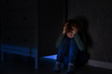 Scared boy hiding behind chest of drawers at night
