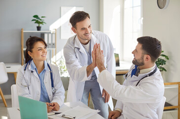 Three cheerful doctors in white coats gathered around table in bright medical office. Smiling physician giving high five to his colleague, team celebrating successful collaboration and achievement.