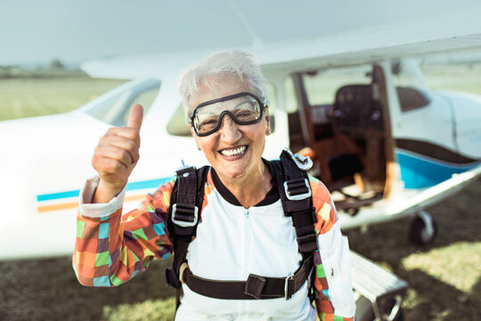 Portrait of a senior woman skydiver smiling and holding a thumbs up with an airplane behind her on a field