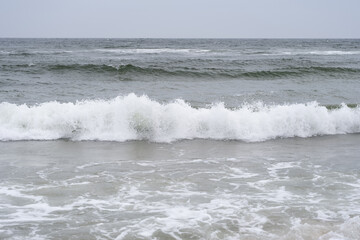 A dramatic sea wave crashes onto the sandy shore under a cloudy sky. The foamy water and dynamic motion capture the raw energy of the ocean.