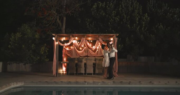 At wedding, diverse couple dancing romantically by pool under beautifully lit gazebo at night