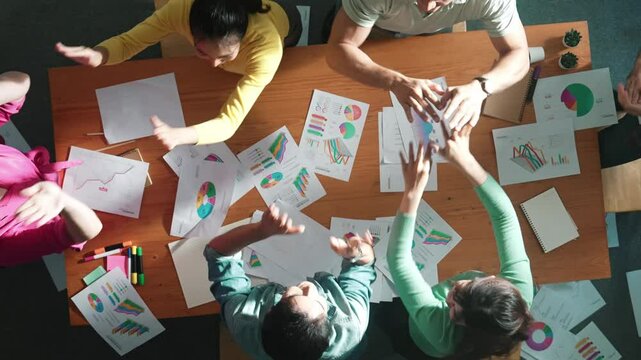 Top view of smart people throw paper and giving high five at meeting table with financial document and stock market analysis paper scattering around. Aerial view of team celebrate project. Symposium.