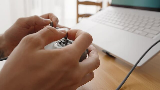 Person Holding and Plugging Cable into FPV Drone Joystick Controller near Laptop on Wooden Desk in Soft Daylight, Close-Up Side View, Manual Operation Concept