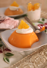 A delicious orange pudding topped with whipped cream and a fresh orange slice, served on a white plate. The dessert has a smooth texture with a bite taken out, set in a cozy dining setting.