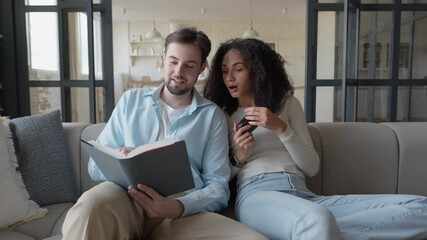 Couple Relaxing On Sofa, Reading Book Together, Reacting With Surprise Interest