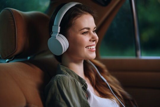 Young woman enjoying music with headphones in a car, smiling happily while relaxing in a green outdoor setting, embodying joy and tranquility.