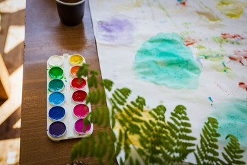 A watercolor palette and painted paper with fern leaf prints on a wooden table.