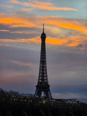 Obraz premium The Eiffel Tower stands silhouetted against a dramatic sunset sky filled with vibrant orange and dark purple clouds, with Parisian building lights glowing softly below.