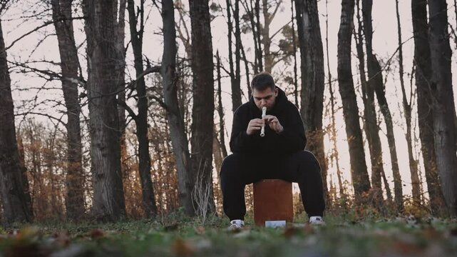 A young man in a black sweatshirt plays on panpipe enchanting melodies in the autumn park.