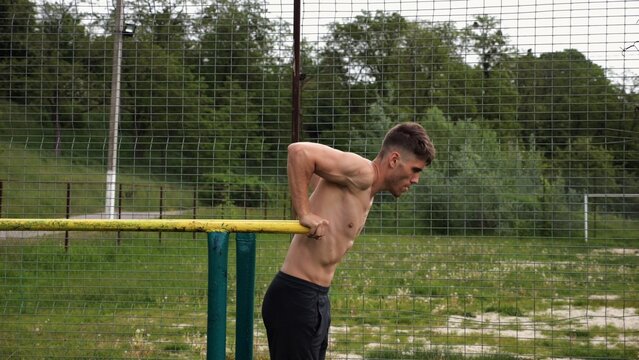 Young sportsman doing push ups on parallel bars at nature. Strong sporty man exercising at sports ground. Male shirtless athlete training hard outdoor. Concept of active sportive lifestyle. Slow mo