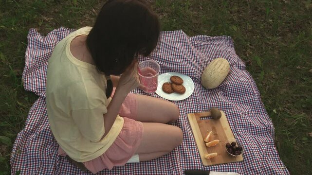 Teenage girl blows through a straw into cup of lemonade, creating bubbles and entertaining herself at picnic. Adolescent girl, funny plays around while sitting on the grass. Top view