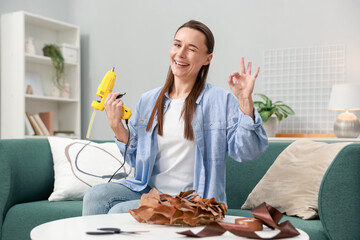 Woman with hot glue gun showing OK gesture at table indoors