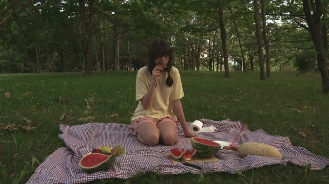Adolescent girl with pigtails bites oatmeal cookies on a summer day. Teenage girl in yellow T-shirt and pink skirt kneels on blanket plaid and eats baked goods during picnic in park. Front view