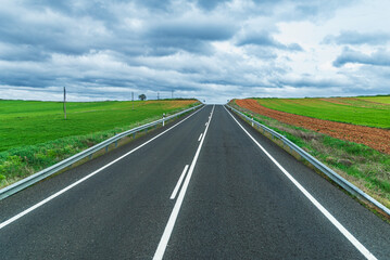 A straight road fading into the distance at the crest of a hill, bordered by green fields and under a stormy sky.