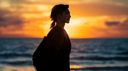 Fototapeta premium Silhouette of Asian woman with ponytail against dramatic orange sunset sky and ocean waves, side profile capturing contemplative peaceful moment at beach during golden hour.
