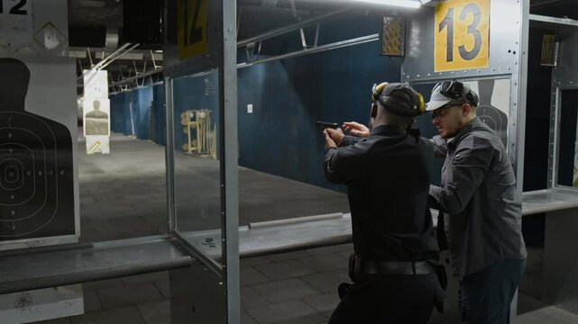 Medium full arc of Caucasian shooting coach and black male security officer in uniform standing together at stall at shooting range, instructor demonstrating how to hold pistol and aim at target