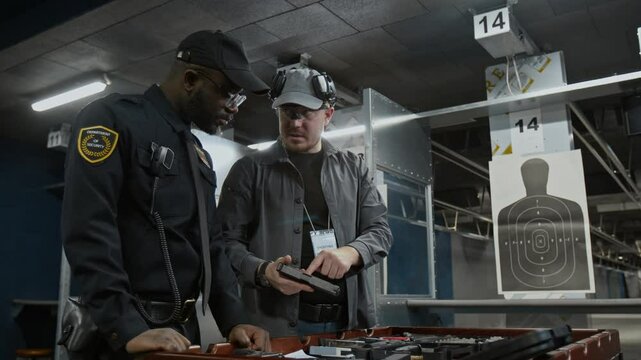 Medium full of young Caucasian male shooting coach teaching trainee African American policeman in black uniform how to load magazine with bullets into handgun during marksmanship course