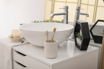 Sink bowl, cup with toothbrushes, soap bars and florarium on chest of drawers in bathroom
