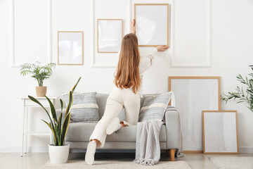 Young woman hanging blank frames on white wall in light living room