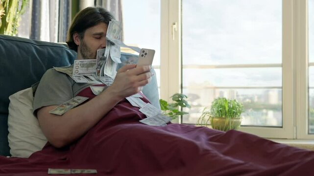 Relaxed man lying in bed using a smartphone as dollar bills fly towards his face, online earnings and financial success. Wealth, passive income, and easy money making from home. Earing money online