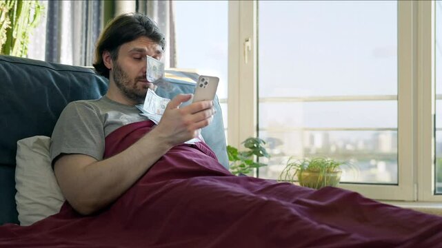 Relaxed man lying in bed using a smartphone as dollar bills fly towards his face, online earnings and financial success. Wealth, passive income, and easy money making from home. Earing money online