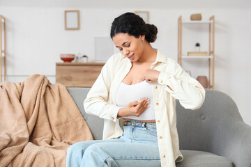 Young African-American woman checking her breast on sofa at home. Cancer awareness concept