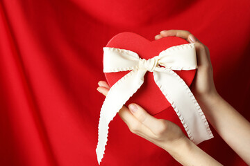 Woman holding heart shaped gift box with beautiful bow against red curtain, closeup. Space for text
