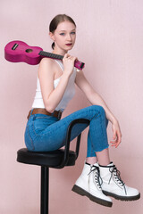 Young female musician sitting on bar stool, holding ukulele by neck and throwing musical instrument over her shoulder, looking at camera. Stylish hipster woman wearing white vest, skinny jeans, boots