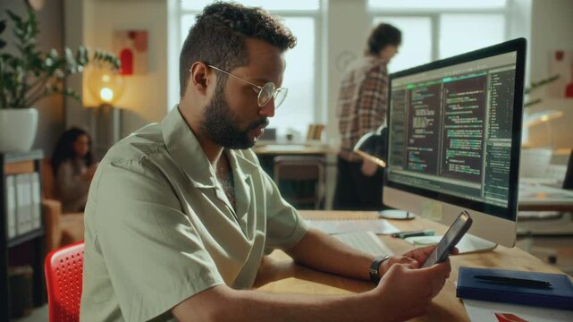 Concentrated male software developer sitting at desk in modern open plan office, checking smartphone as code runs on computer monitor, colleagues working in background