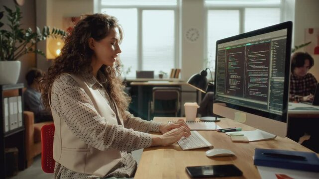 Focused female software developer coding on computer at desk, typing on keyboard and analyzing code on monitor in modern open place office with colleagues working in background