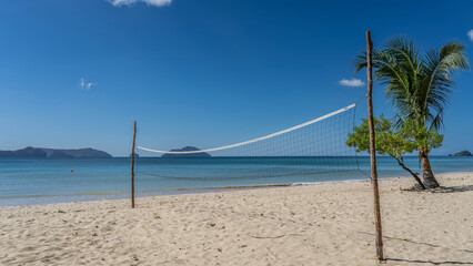 Beach volleyball net on a tropical sandy sandy coast of the sea. A calm turquoise ocean. Silhouettes of mountains in the distance. Blue sky, clouds. Palm tree by the water. Philippines. Palawan. 