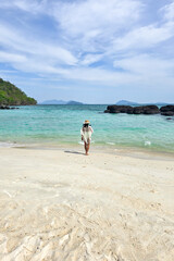 Serenity envelops the beautiful shores of Koh Kham Island, where a lone figure strolls along the soft, white sand, embracing the turquoise waves under a clear blue sky in Thailand