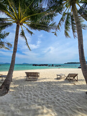 Warm sunlight filters through tropical palms, casting shadows on the white sandy beach of Koh Kham Thailand. Loungers invite relaxation next to the tranquil turquoise waters