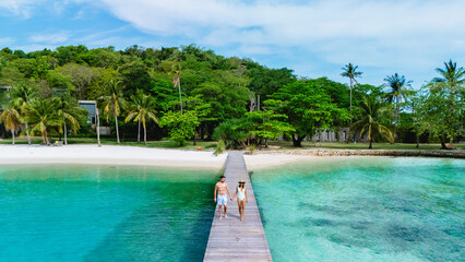 A romantic couple strolls hand in hand along a wooden pier, surrounded by the crystal-clear waters of Koh Kham Island Thailand. Lush greenery and a pristine beach create a tranquil atmosphere.