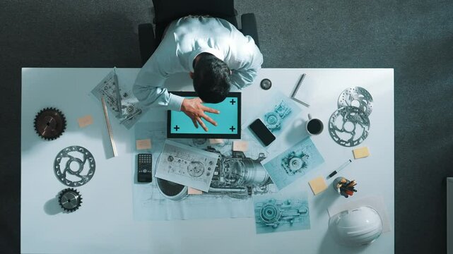 Top view of engineer looking at phone while working on tablet with green screen at meeting table with turbine engine or jet engine sketch design and various metal gear scatter around. Alimentation.