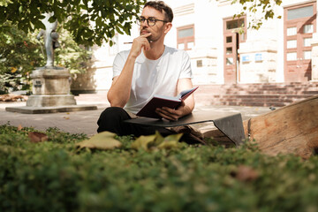 Thoughtful man sitting outdoors on a bench, holding a notebook, surrounded by greenery and urban architecture. He appears lost in thought, capturing the essence of inspiration and creativity.