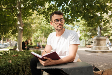 Fototapeta premium A young man sits on a park bench, writing in a red journal on a sunny day. Surrounded by greenery, he looks thoughtful and relaxed in this serene outdoor setting.