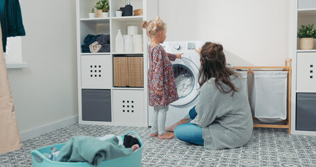 A smiling mom and her little daughter spend time in the laundry room, doing chores during maternity...