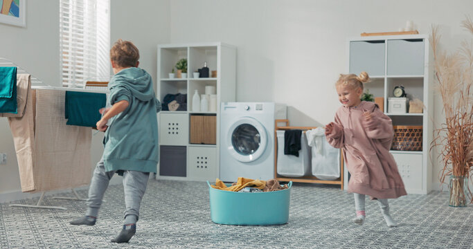 Happy siblings are fooling around in the laundry area, wrapped in warm sweatshirts fresh from the dryer, running between the clothes bowl and the bathroom washing machine.