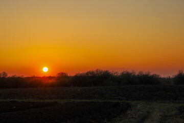 The photograph shows a sunset over a field. The sun sets behind a line of trees, illuminating the sky with bright orange and yellow colors.