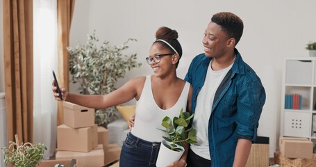 A young couple just moved in. She stands holding a potted plant, he grabs his phone. They take a selfie in their new space, laugh, and scroll through the photos to choose the best one. © ABCreative