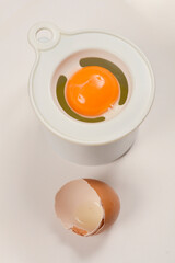 A white plastic egg separator with a bright orange yolk inside is placed above a cracked eggshell on a white surface